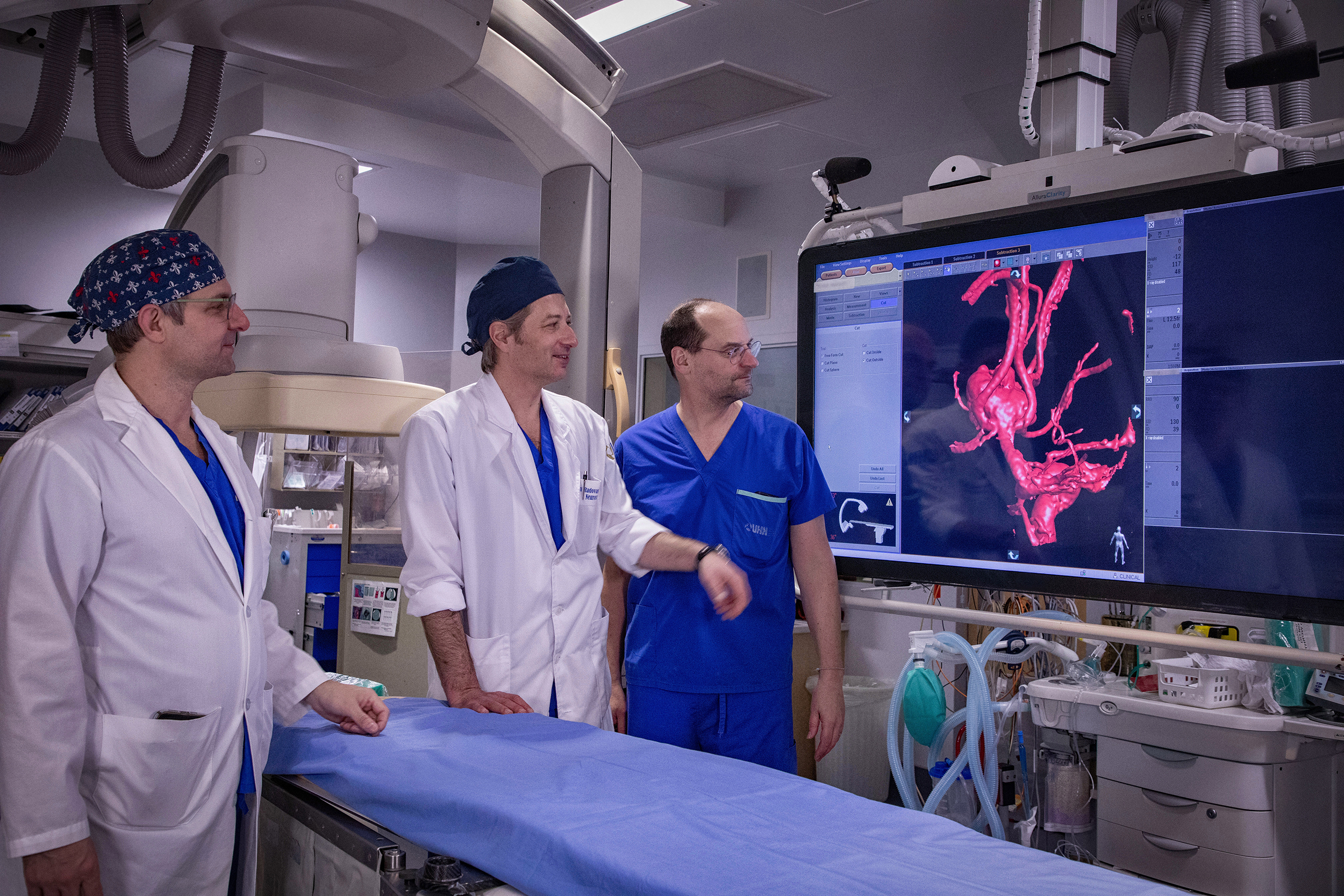 Three members of the Toronto Neurovascular group standing at the operating table looking at a screen that is displaying medical images.